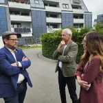 Bundestagsabgeordneter Lars Rohwer im Gespräch mit FASA‑AG‑Geschäftsführer Ulrich Hintzen und einer Kollegin vor einem modernen Mehrfamilienhaus mit solaraktiver Fassadenverkleidung.