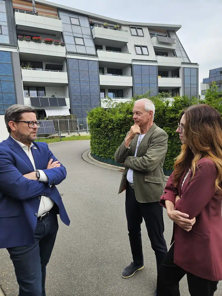 Bundestagsabgeordneter Lars Rohwer im Gespräch mit FASA‑AG‑Geschäftsführer Ulrich Hintzen und einer Kollegin vor einem modernen Mehrfamilienhaus mit solaraktiver Fassadenverkleidung.