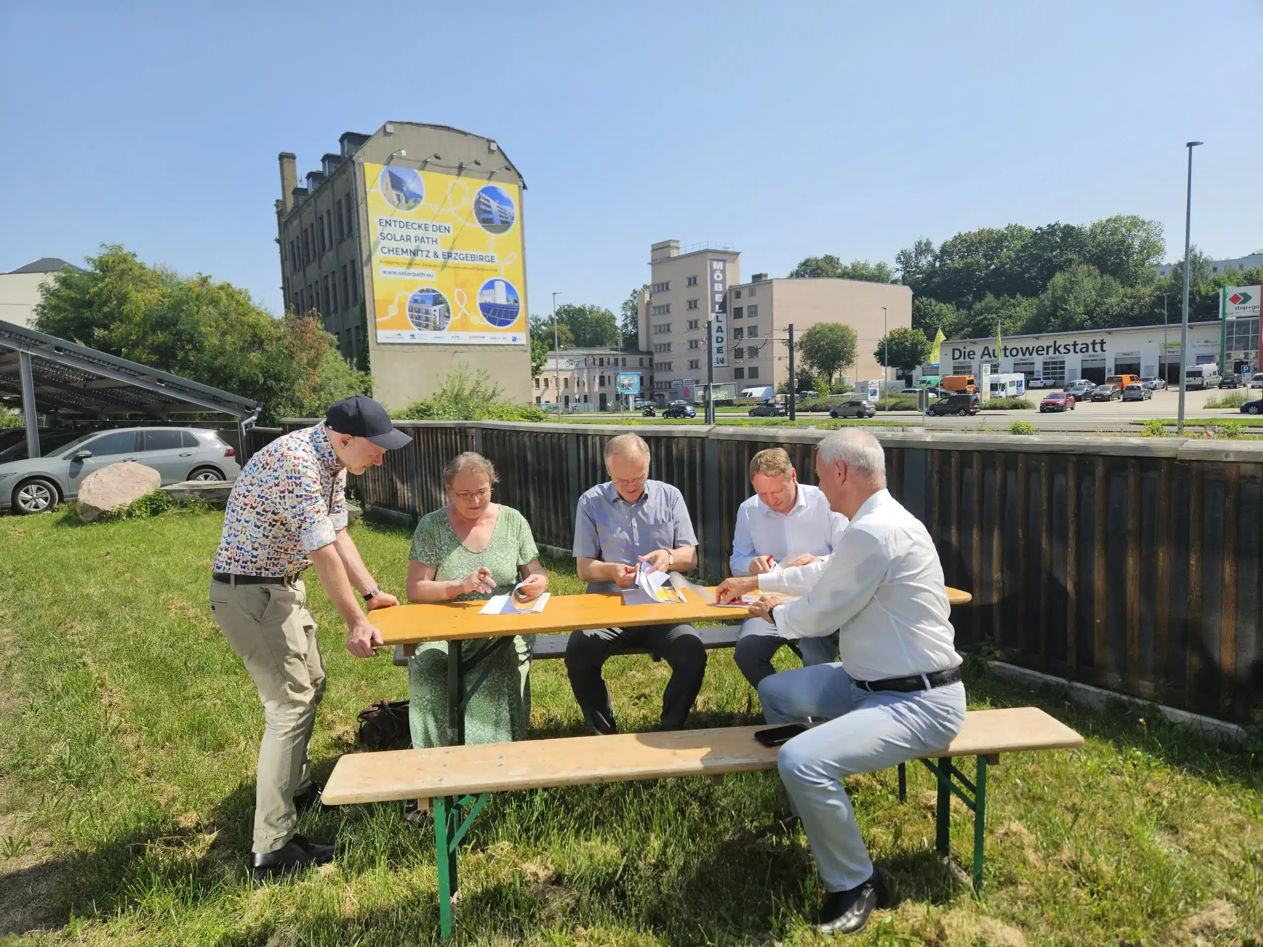 Bei sommerlichem Wetter unterzeichnen mehrere Vertreterinnen und Vertreter des Solarpfads Chemnitz-Erzgebirge an einem Biertisch im Freien eine Kooperationsvereinbarung; im Hintergrund sind ein großes Solarpfad-Banner an einer Fassade, eine überdachte PV-Parkplatzanlage sowie umliegende Gewerbebauten zu sehen.