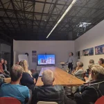 Zum Jubiläum „35 Jahre FASA“ sitzen zahlreiche Gäste im Showroom der FASA AG um einen langen Holztisch und verfolgen einen Vortrag, bei dem auf einem großen Bildschirm Bauaufnahmen eines Aktivsonnenhaus-Projekts gezeigt werden; vorne steht ein Referent am Laptop, auf den Wänden sind weitere Projektfotos und Auszeichnungen des Unternehmens zu sehen.