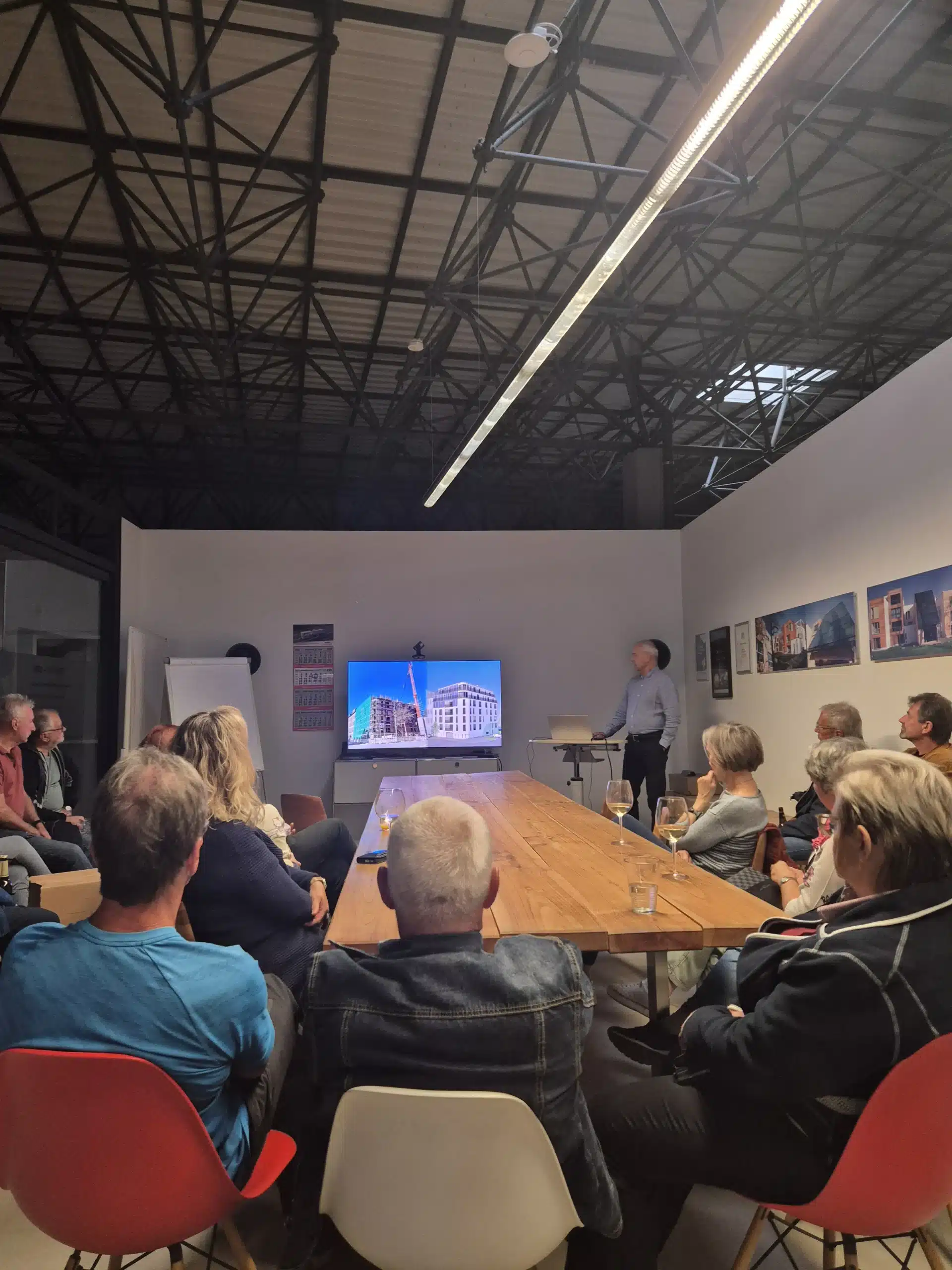 Zum Jubiläum „35 Jahre FASA“ sitzen zahlreiche Gäste im Showroom der FASA AG um einen langen Holztisch und verfolgen einen Vortrag, bei dem auf einem großen Bildschirm Bauaufnahmen eines Aktivsonnenhaus-Projekts gezeigt werden; vorne steht ein Referent am Laptop, auf den Wänden sind weitere Projektfotos und Auszeichnungen des Unternehmens zu sehen.