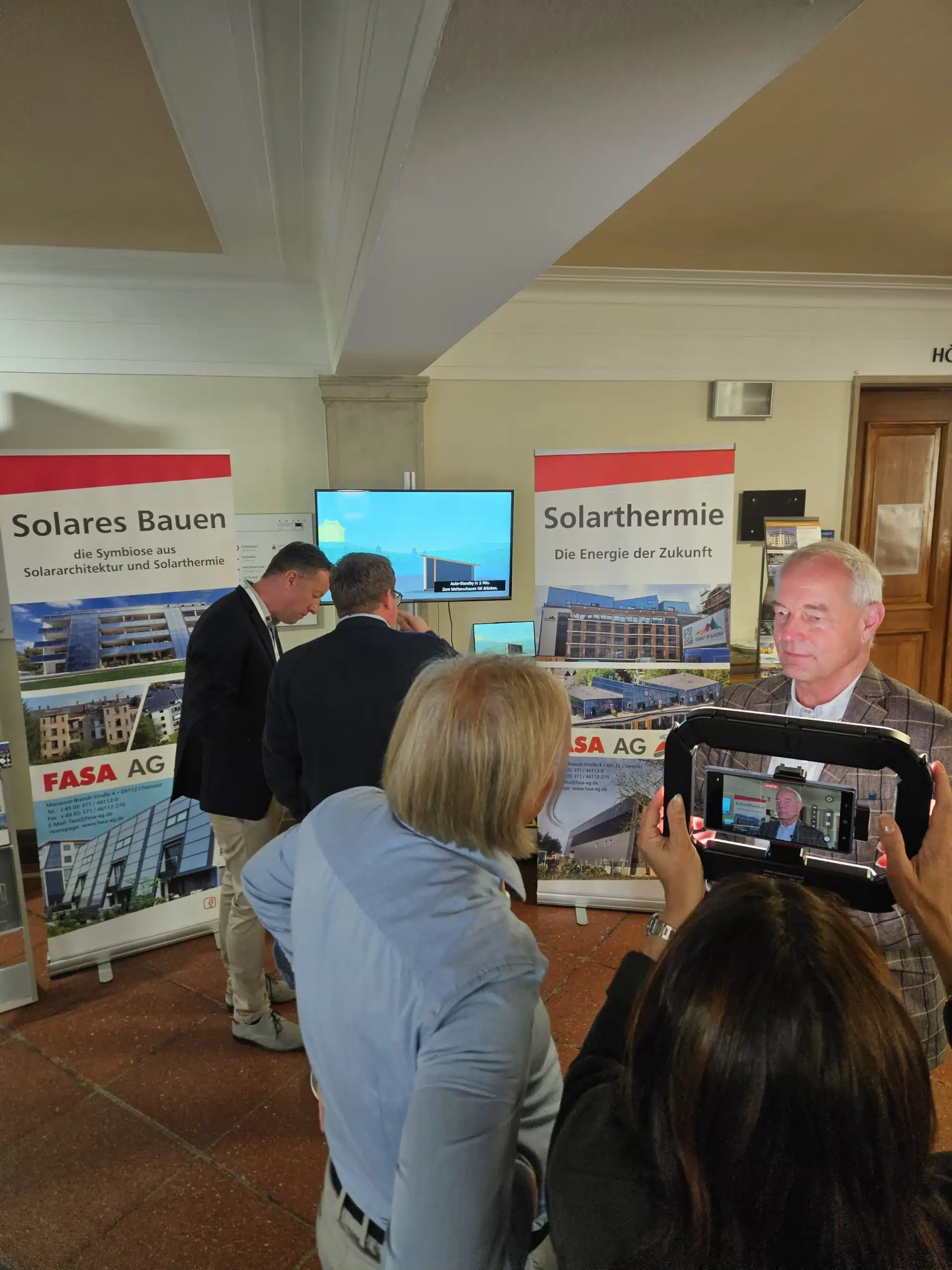 Beim Ostdeutschen Energieforum (ODEF) in Leipzig präsentiert die FASA AG ihren Stand zum Thema solares Bauen und Solarthermie: Vor zwei Roll-ups mit der Aufschrift ‚Solares Bauen‘ und ‚Solarthermie – Die Energie der Zukunft‘ finden Fachgespräche statt, während im Vordergrund ein Interview mit Geschäftsführer Ulrich Hintzen als Vertreter der FASA AG per Tablet-Kamera aufgezeichnet wird.