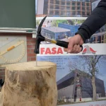 Beim „Tag der Bildung“ demonstriert ein Teilnehmer am Stand der FASA AG handwerkliches Geschick: Vor einem FASA-Roll-up mit Fotos von Aktivsonnenhaus-Projekten wird mit einem Latthammer ein Nagel in einen massiven Holzstamm geschlagen, im Hintergrund sind Tafel und Bauzeichnungen zu sehen.