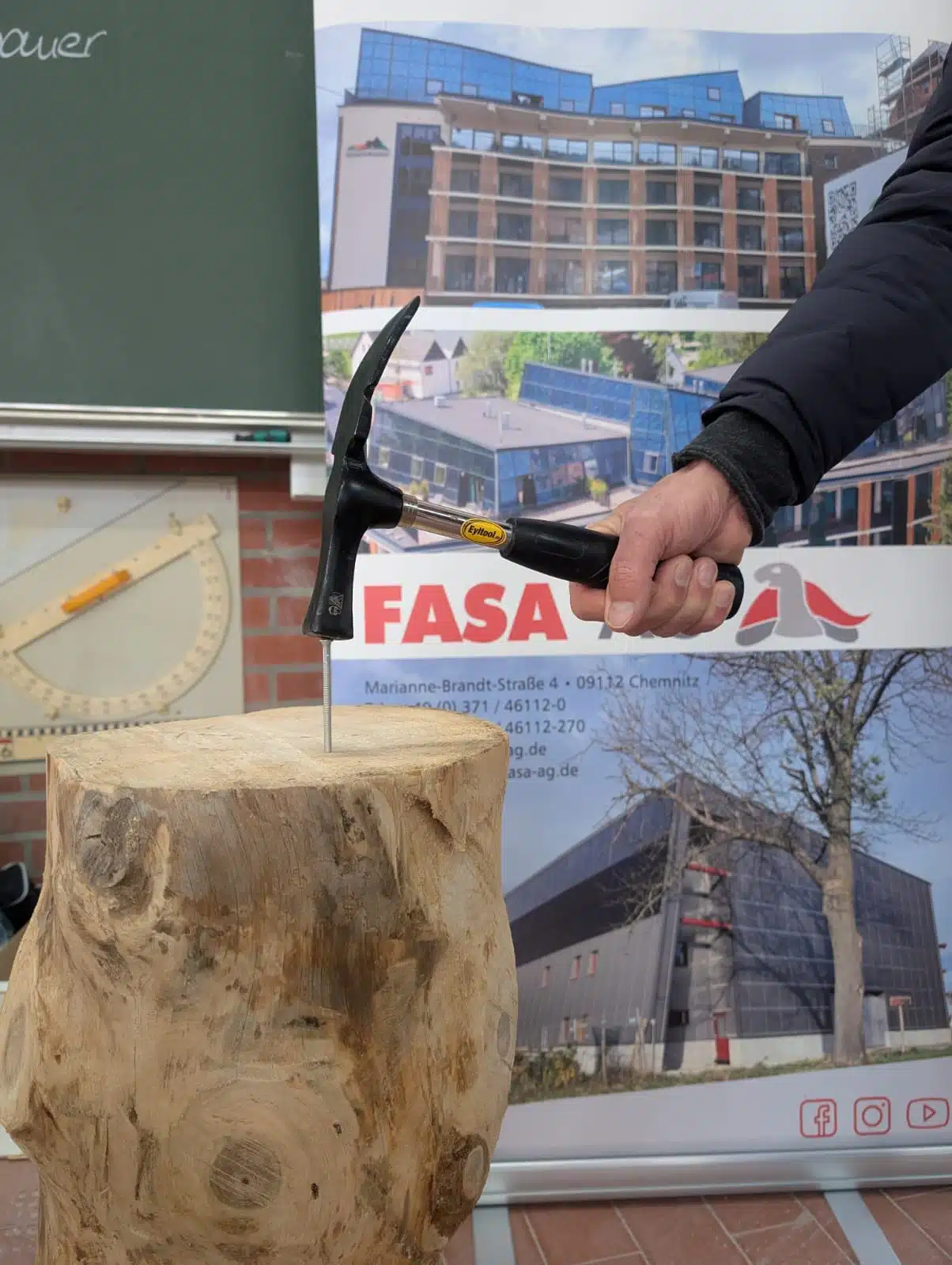 Beim „Tag der Bildung“ demonstriert ein Teilnehmer am Stand der FASA AG handwerkliches Geschick: Vor einem FASA-Roll-up mit Fotos von Aktivsonnenhaus-Projekten wird mit einem Latthammer ein Nagel in einen massiven Holzstamm geschlagen, im Hintergrund sind Tafel und Bauzeichnungen zu sehen.