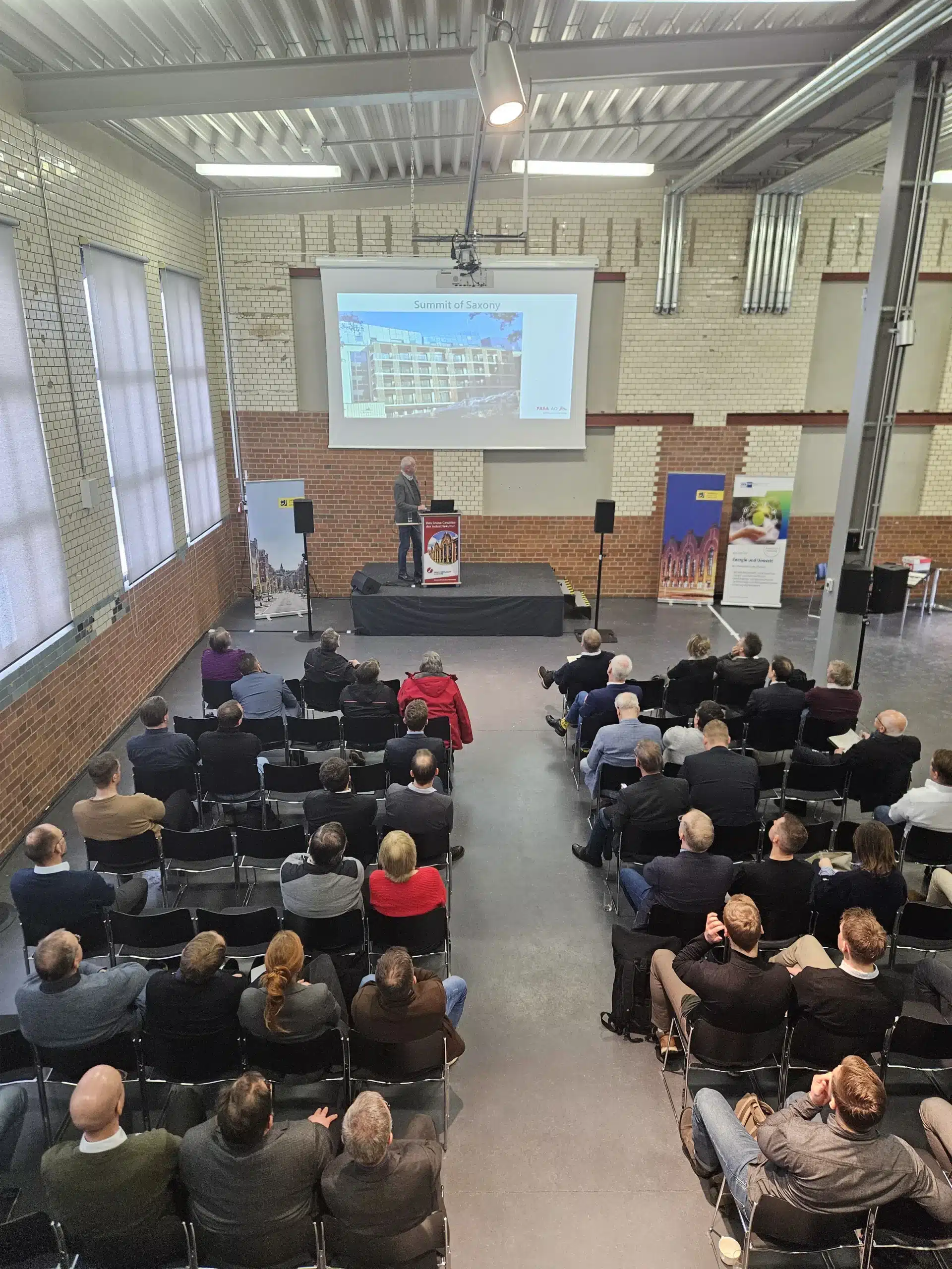 Beim 4. Energiedialog Chemnitz am 27. Januar 2026 präsentiert ein Referent auf einer Bühne im Industrieloft vor voll besetztem Saal das Projekt „Summit of Saxony“: Auf der Leinwand ist ein Aktivsonnenhaus-Gebäude der FASA AG zu sehen, während zahlreiche Fachgäste in Reihen sitzen und den Vortrag zur zukünftigen Energieversorgung in Sachsen verfolgen.