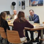 Beim Besuch des sächsischen Landtagspräsidenten sitzen mehrere Personen mit ihm an einem rechteckigen Holztisch im Besprechungsraum der FASA AG, vor ihnen stehen Wasser- und Saftflaschen, Kaffee und Notizen; im Hintergrund sind ein Laptop, Auszeichnungen und Fotos von Aktivsonnenhäusern an der Wand zu sehen, was den Rahmen für einen gegenseitigen Impuls- und Austauschbesuch bildet.