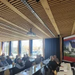 Im Seminarraum der Hochschule Weihenstephan-Triesdorf verfolgt ein vollbesetztes Plenum die Jahreshauptversammlung des Sonnenhaus-Instituts: Vorne rechts präsentiert ein Referent vor einem großen Banner „Das Sonnenhaus“ mit solarthermischem Gebäude, während die Teilnehmenden an langen Tischen mit Getränken und Unterlagen sitzen; im Hintergrund informieren mehrere Roll-ups über Projekte und Inhalte rund um erneuerbare Energien.