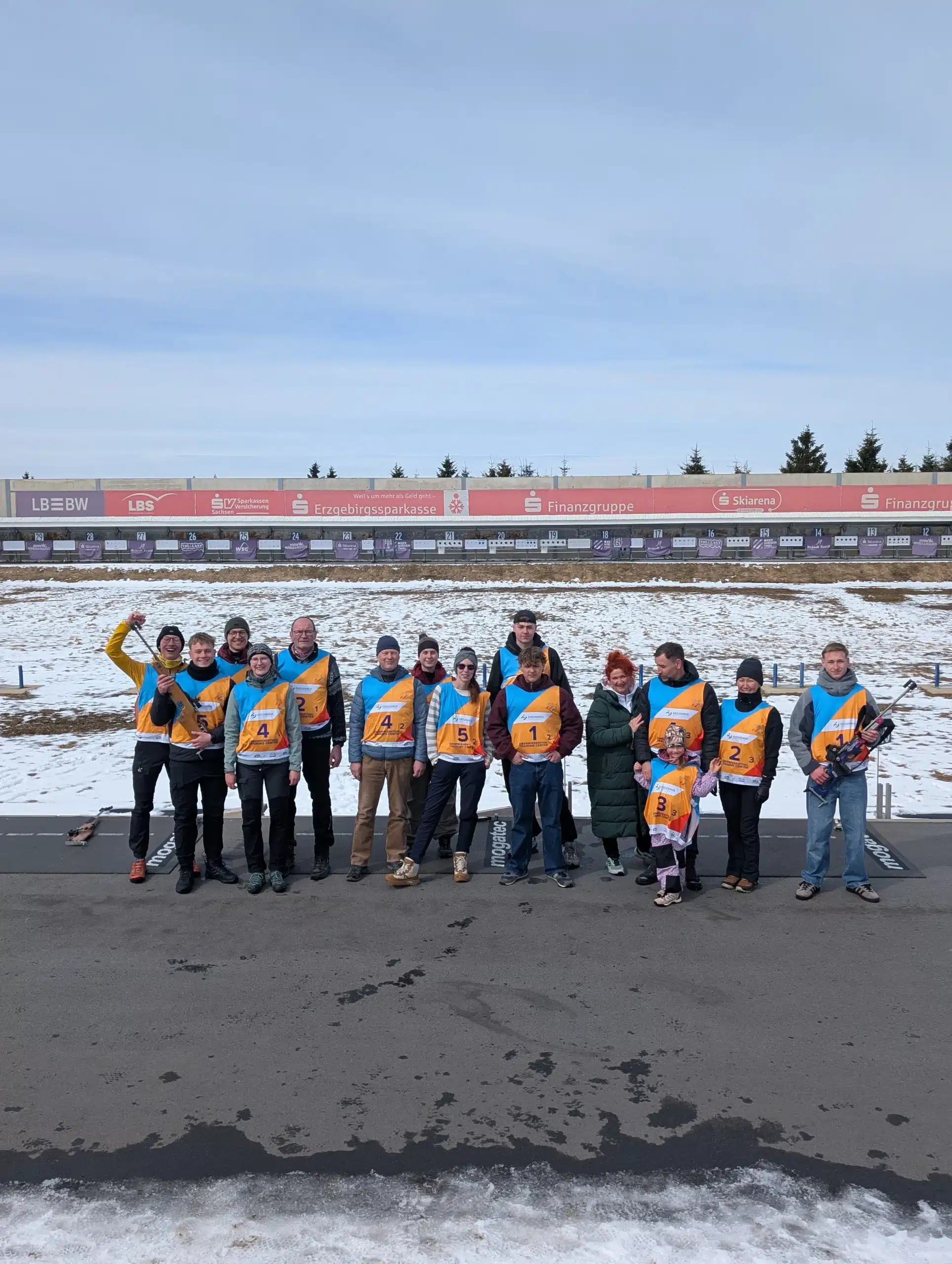 Gruppenfoto vom FASA-Sporttag in Oberwiesenthal: Eine größere Gruppe von Mitarbeitenden und Angehörigen steht auf der teilweise verschneiten Biathlonanlage, alle tragen orange-blaue Startnummernwesten, im Hintergrund sind die beschilderten Schießstände sowie Bandenwerbung der Sparkasse und weiterer Partner zu sehen.