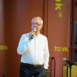 Auf dem Foto spricht der Geschäftsführer Ulrich Hintzen beim Jubiläum der Jenni Energietechnik AG, langjähriger Partner der FASA AG, vor einem großen, orangefarbenen Jenni-Wärmespeicher mit der gelben Aufschrift „700 kWh“. Im Vordergrund steht er in weißem Hemd mit Mikrofon in der Hand neben einem Rednerpult mit Laptop, im Hintergrund sind Rohrleitungen und weitere Anlagenteile der Wärmetechnik zu sehen.