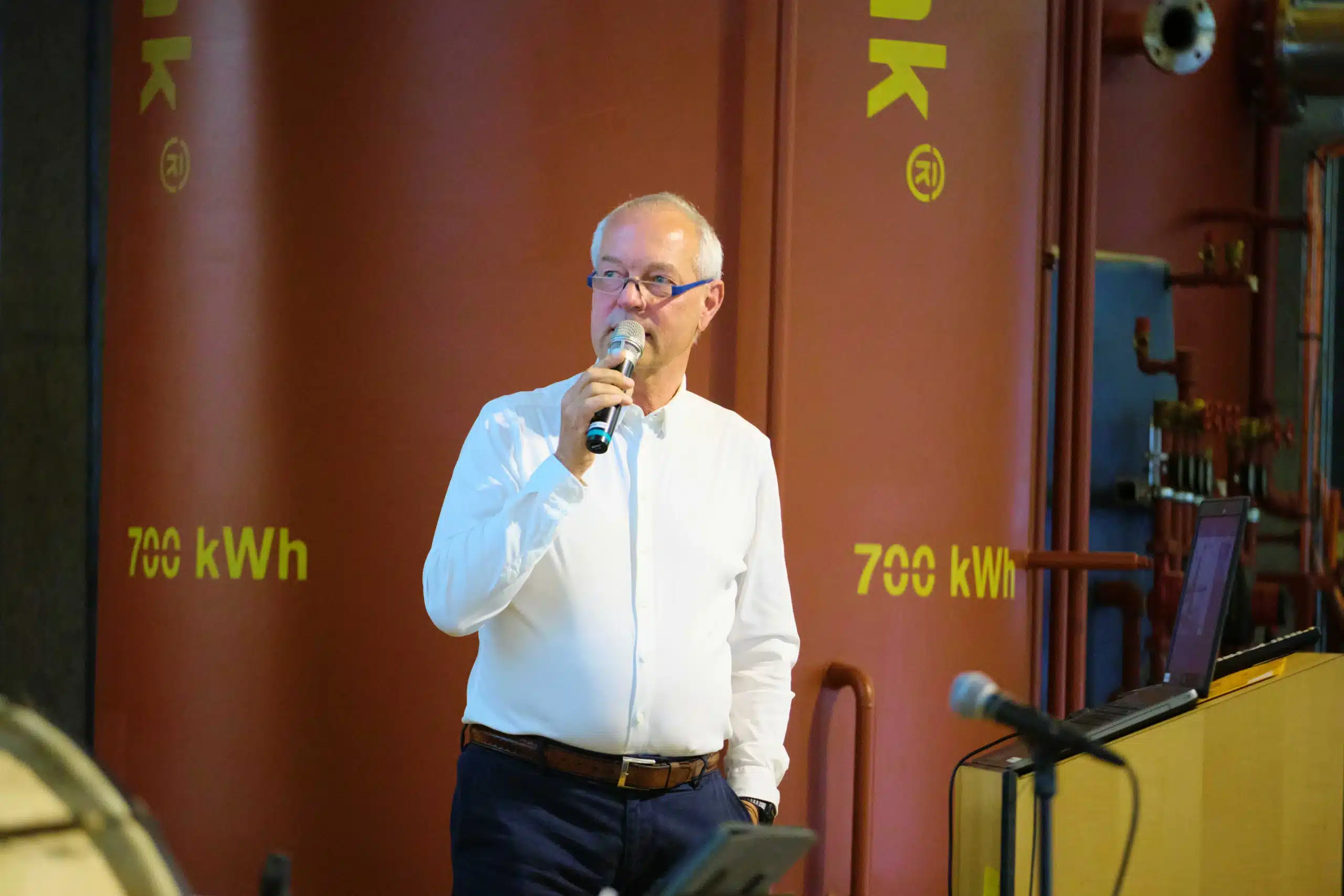 Auf dem Foto spricht der Geschäftsführer Ulrich Hintzen beim Jubiläum der Jenni Energietechnik AG, langjähriger Partner der FASA AG, vor einem großen, orangefarbenen Jenni-Wärmespeicher mit der gelben Aufschrift „700 kWh“. Im Vordergrund steht er in weißem Hemd mit Mikrofon in der Hand neben einem Rednerpult mit Laptop, im Hintergrund sind Rohrleitungen und weitere Anlagenteile der Wärmetechnik zu sehen.