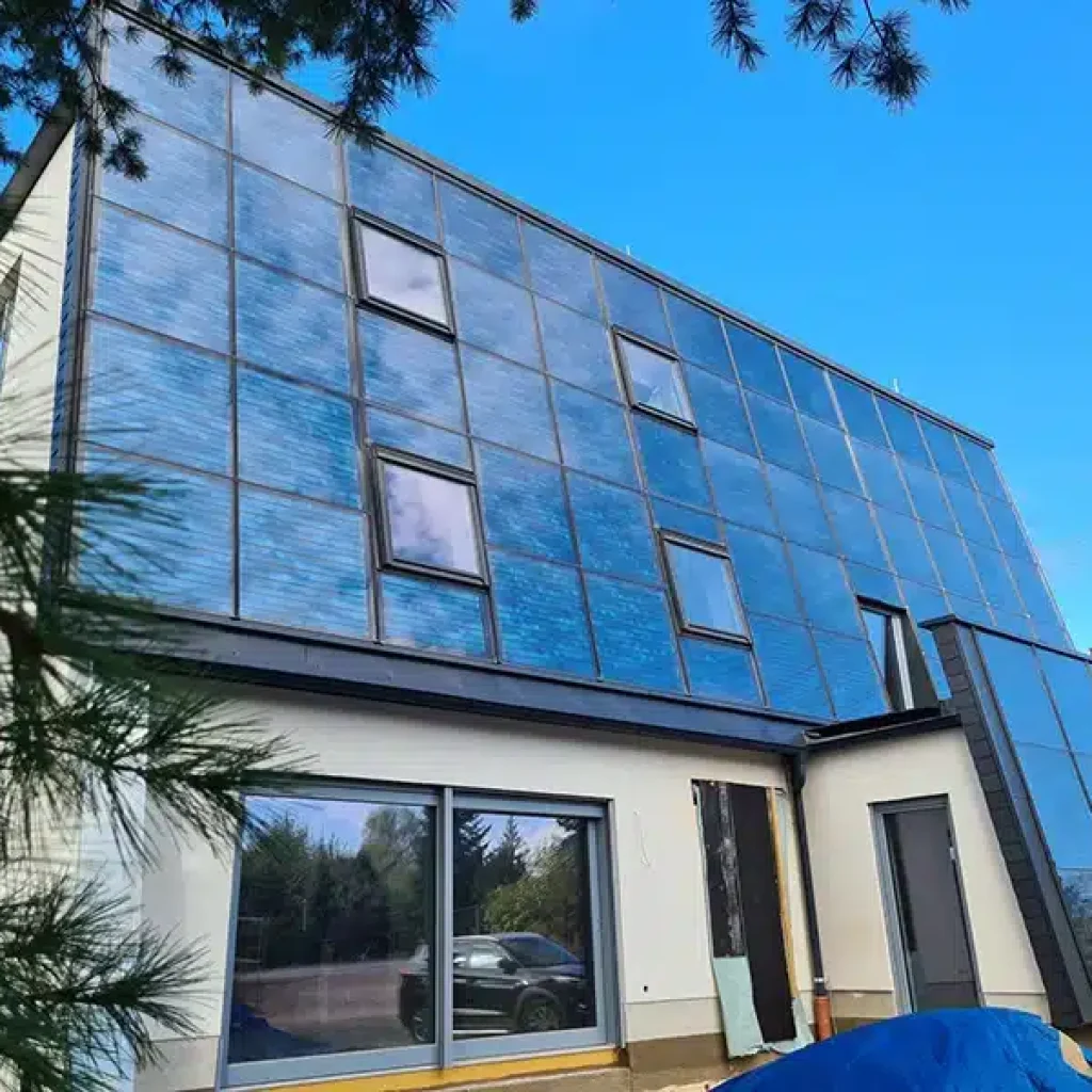 Moderne Solarvilla Schlossblick der FASA AG mit großflächiger, blauer Solar-Glasfassade und integrierten Fenstern, aufgenommen aus der Froschperspektive bei blauem Himmel; im Erdgeschoss große Panoramafenster, im Vordergrund ein abgedeckter Materialhaufen und seitlich Bäume am Bildrand.